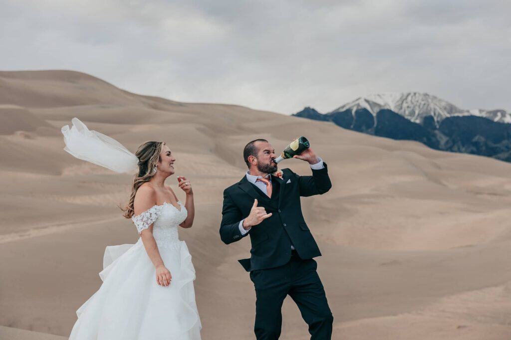 a groom drinking from a champagne bottle while his bride laughs at him at great sand dunes national park